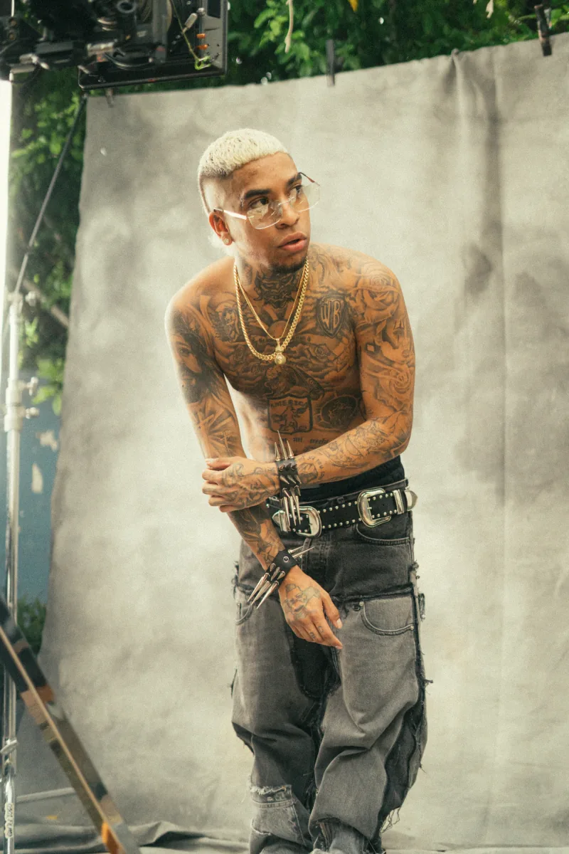 Artista con el torso tatuado y pelo platinado frente a un lienzo gris de estudio con humo en el aire